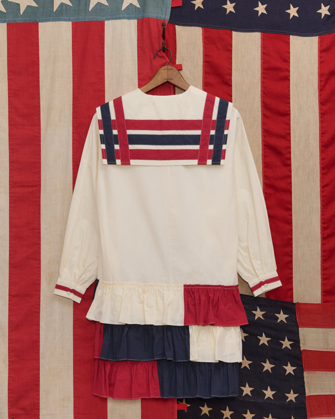 The Centennial Dress - Multi features red, white, and blue stripes with ruffles, displayed against an American flag backdrop—evoking classic Fourth of July charm and vintage Liberty dress styles.