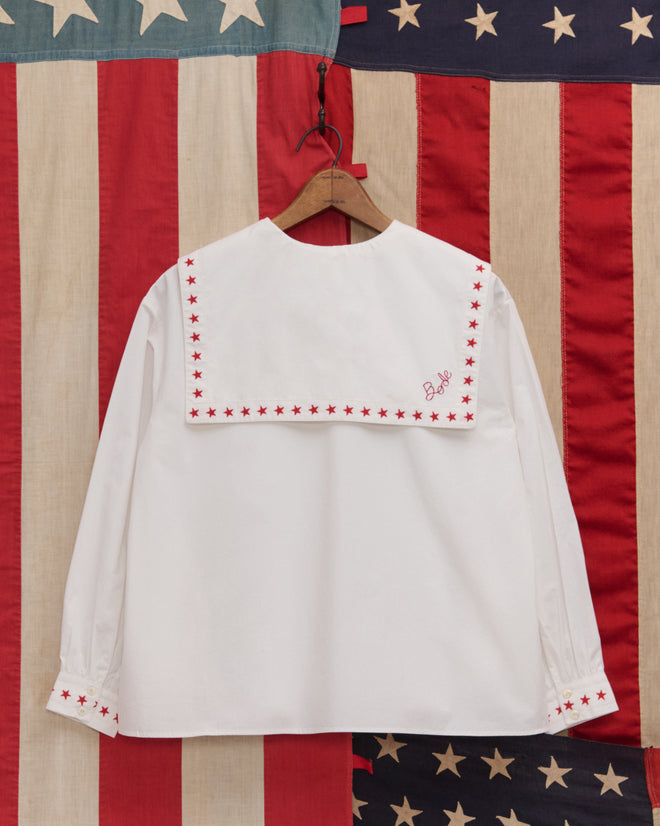 Red Star Blouse: White sailor-style top with red star trim, displayed on a wooden hanger against an American flag backdrop—ideal for Fourth of July or Liberty-themed costumes.