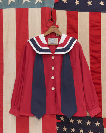 The Centennial Blouse, a red sailor-style top with navy and white collar details, hangs on a wooden hanger against an American flag backdrop.