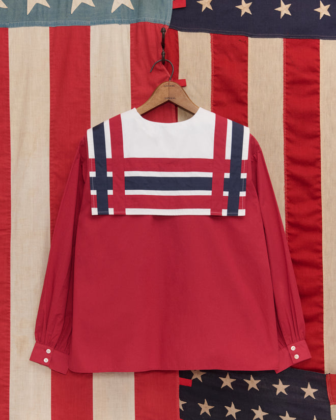 The Centennial Blouse, a red long-sleeve top with a nautical striped collar inspired by antique Liberty blouses, is displayed against vintage American flag fabric.