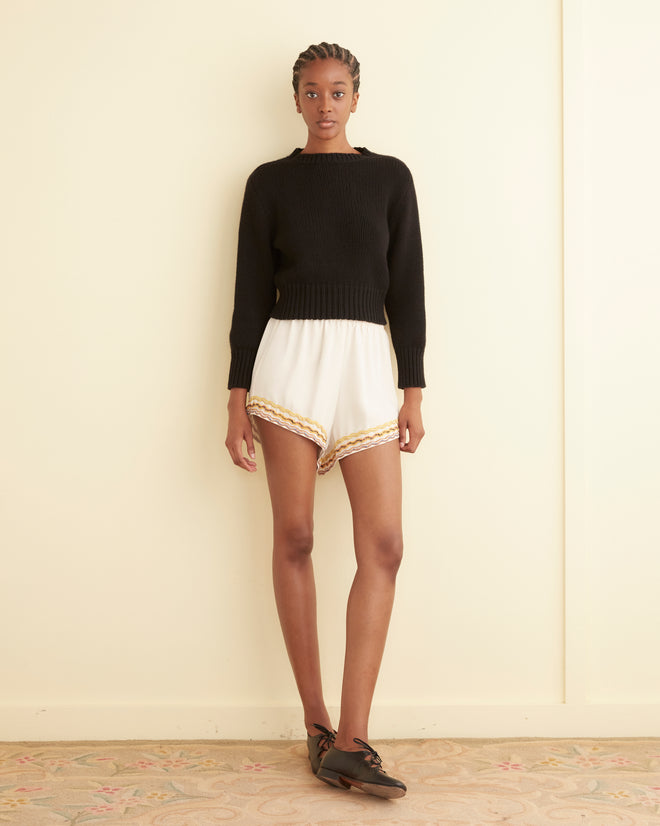 A person is indoors wearing a black sweater and Rickrack Hem Tap Shorts, which are white with gold trim and embroidered rickrack stripes. They complete the look with black shoes, wearing a neutral expression.