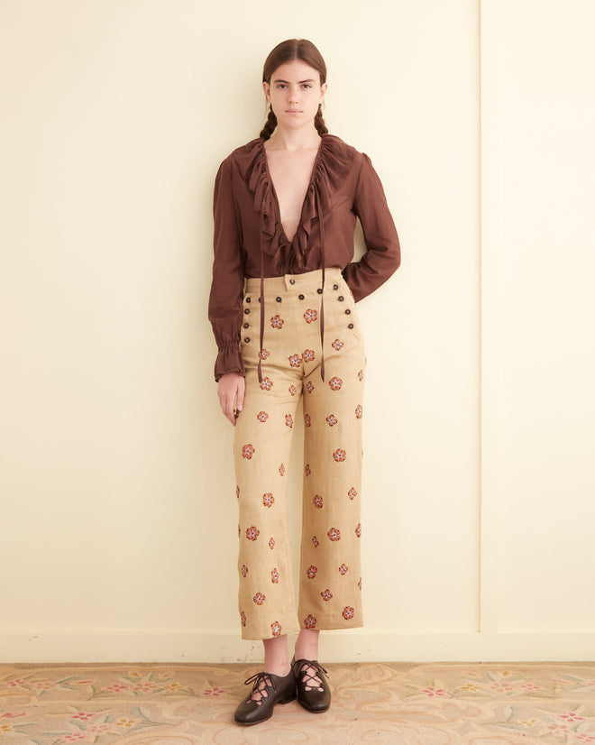 Woman standing against a light-colored wall wearing a brown blouse with ruffles, Embroidered Waxflower Trousers featuring a floral pattern, and black shoes with laces. She has long brown hair styled in pigtails, accentuating her look with an Edwardian women's coat adorned with hand-embroidered flowers.