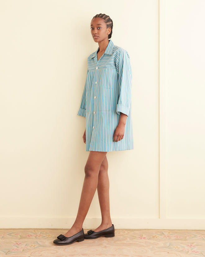 A person stands against an off-white wall, wearing the Shore Stripe Quincy Dress with textured black and white stripes, paired with black flat shoes.