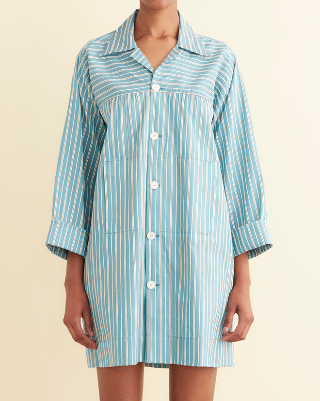 A person stands against an off-white wall, wearing the Shore Stripe Quincy Dress with textured black and white stripes, paired with black flat shoes.