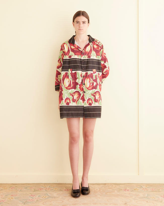 Wearing an oversized Hibiscus Stripe Dress, a woman stands against a light wall on a patterned carpet, evoking the style of a 1960s house coat.