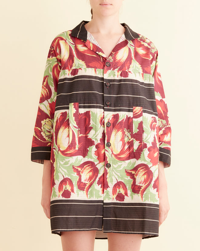 Wearing an oversized Hibiscus Stripe Dress, a woman stands against a light wall on a patterned carpet, evoking the style of a 1960s house coat.