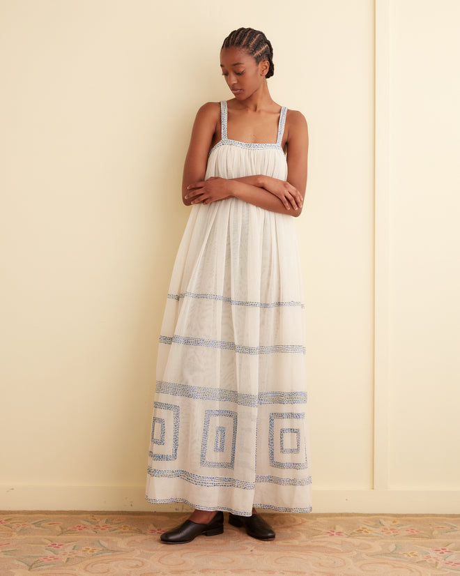 A person stands with arms crossed wearing the Tile Stitch Dress, a long, 1930s inspired design adorned with geometric blue embroidery. They are facing slightly sideways in front of a beige wall.