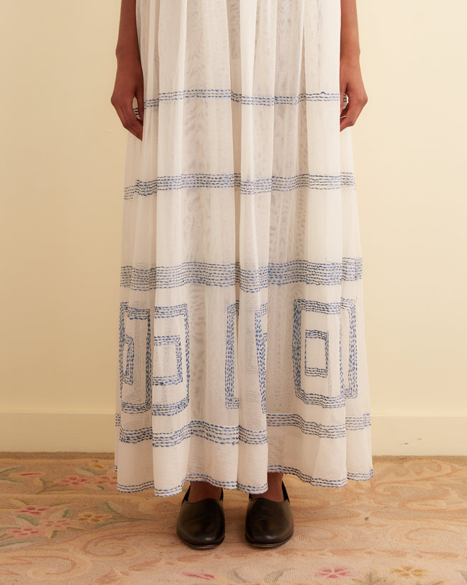 A person stands with arms crossed wearing the Tile Stitch Dress, a long, 1930s inspired design adorned with geometric blue embroidery. They are facing slightly sideways in front of a beige wall.