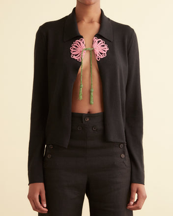 A person wears the Beaded Abilene Cardigan, a black open-front jacket styled over a 1920s-inspired sweater with a pink and green chest tie detail.