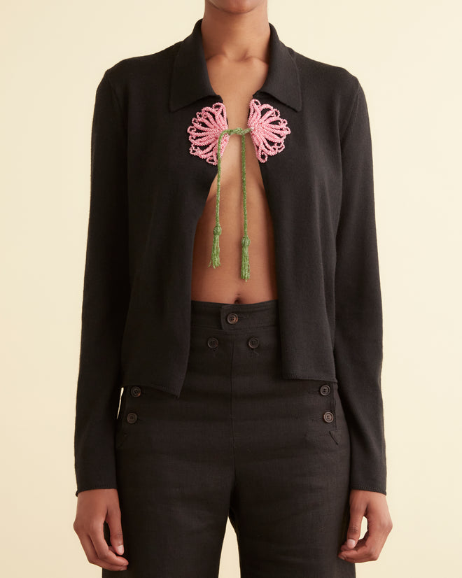 A person wears the Beaded Abilene Cardigan, a black open-front jacket styled over a 1920s-inspired sweater with a pink and green chest tie detail.
