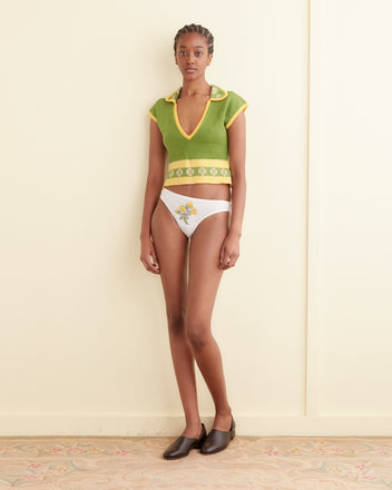 A person stands against a plain wall wearing the green Wellfleet Jacquard Top with yellow trim and white floral-design underwear, along with brown flat shoes. The top features subtle geometric detailing that adds a touch of sophistication to the casual ensemble.