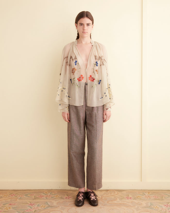 Person standing indoors against a beige wall, wearing an Embroidered Flower Study Shirt, gray trousers, and dark shoes.