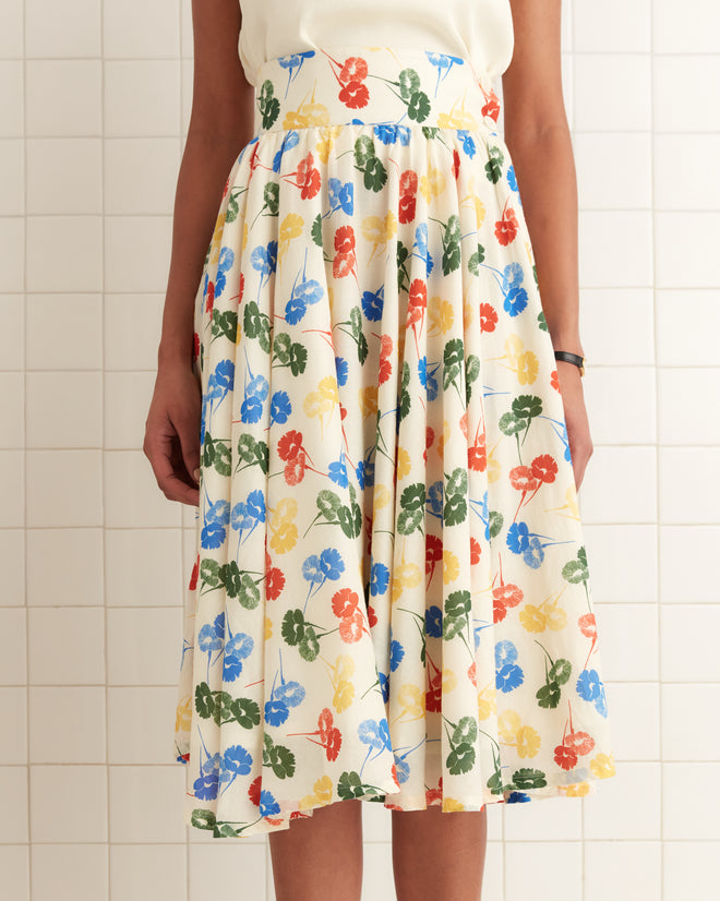 A person stands against a tiled wall wearing the Flower Stamp Skirt, a vibrant white skirt with a colorful floral pattern, made from 100% cotton.