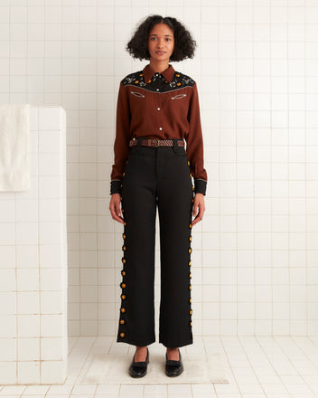 In a tiled room, someone wears a brown shirt paired with Abilene Trousers, featuring black fabric adorned with studded felt flowers.