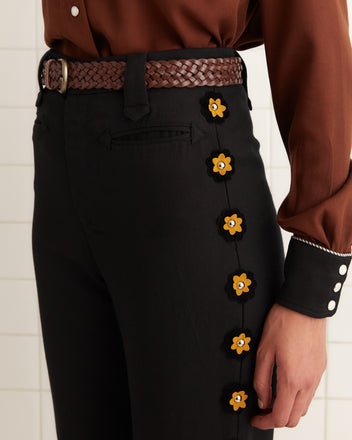 A person wearing Abilene Trousers in lightweight wool twill black fabric featuring yellow floral details, paired with a brown braided belt and a brown shirt adorned with white buttons.