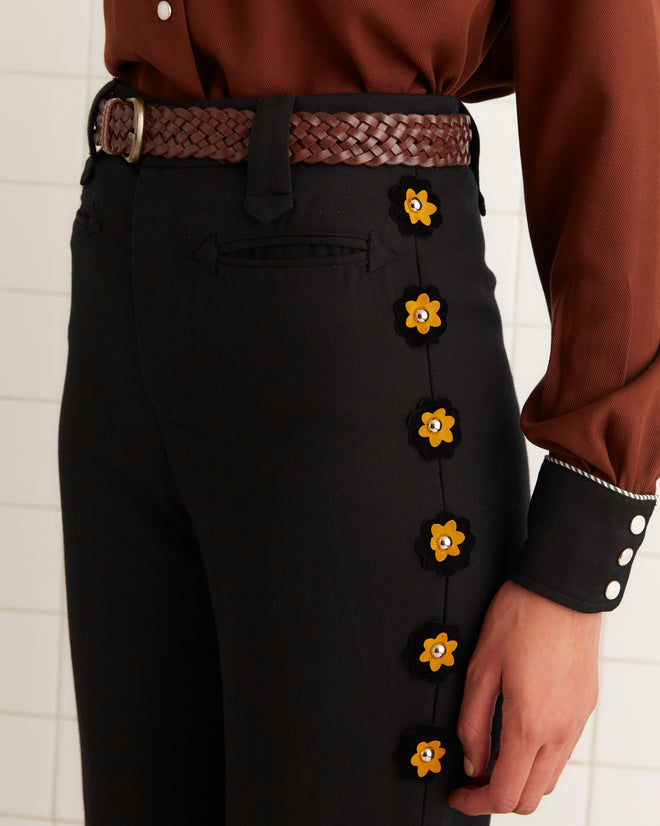 Someone wearing the Abilene Trousers, featuring black pants with studded felt flowers on the side and a braided belt, captures the charm of Western TV show attire.