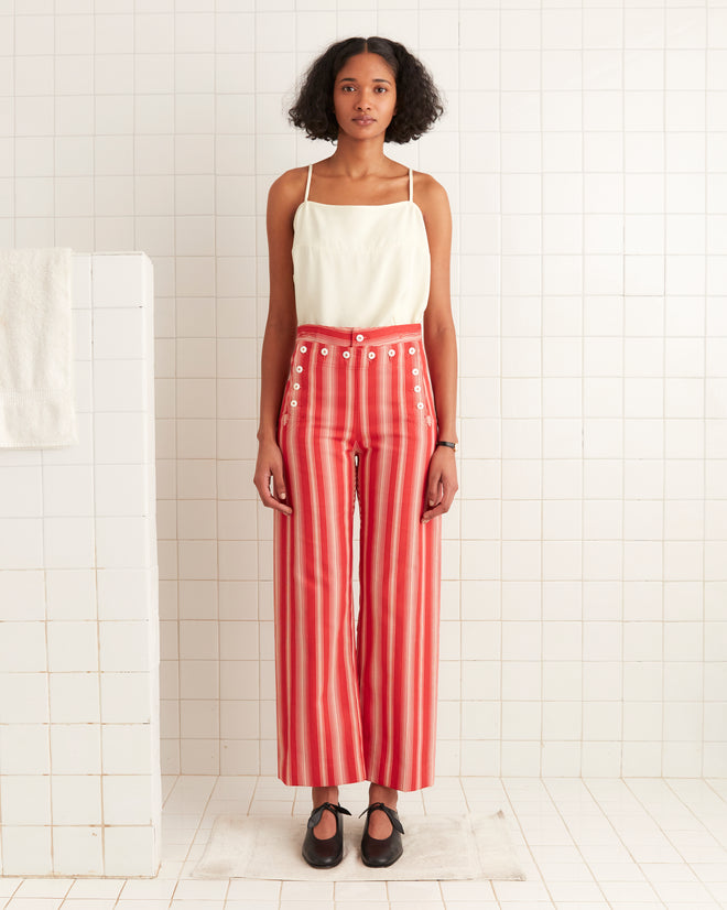 A woman in a white top and Parasol Stripe Sailor Trousers with striking red stripes and pearl buttons stands in a tiled room, leaning gracefully against a small wall.