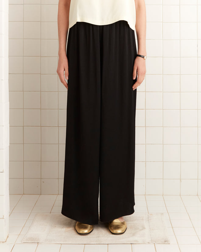 A person stands on a bath mat against a tiled wall, wearing the Roy Pants - Black, inspired by 1920s fashion, paired with gold shoes and a watch.