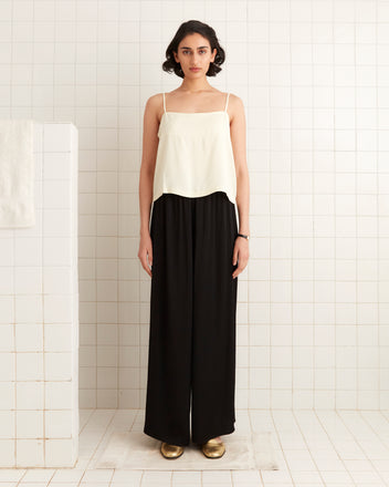 A person stands against a tiled wall, embracing 1920s fashion with a white top, Roy Pants - Black, and gold shoes.