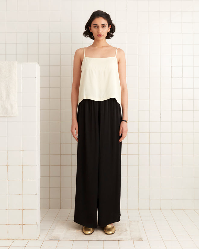 A person stands against a tiled wall, embracing 1920s fashion with a white top, Roy Pants - Black, and gold shoes.