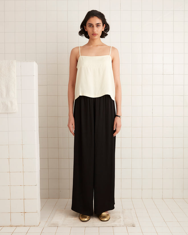 Woman standing in a tiled room, wearing a white camisole, gold shoes, and Roy Pants - Black: wide-leg women’s pants inspired by 1920s fashion.