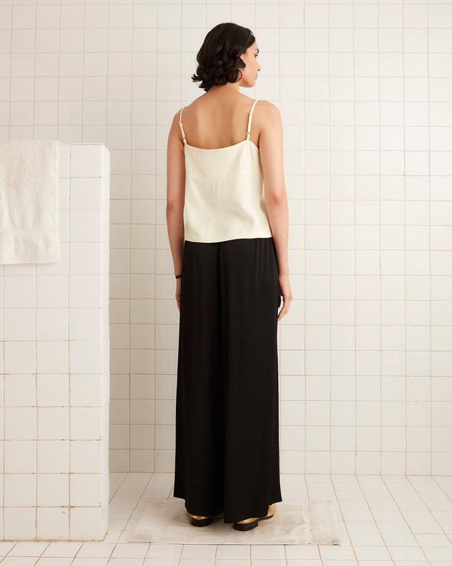 Woman with short dark hair wears a cream camisole and Roy Pants - Black, standing in a tiled bathroom with a look that subtly channels 1920s elegance.