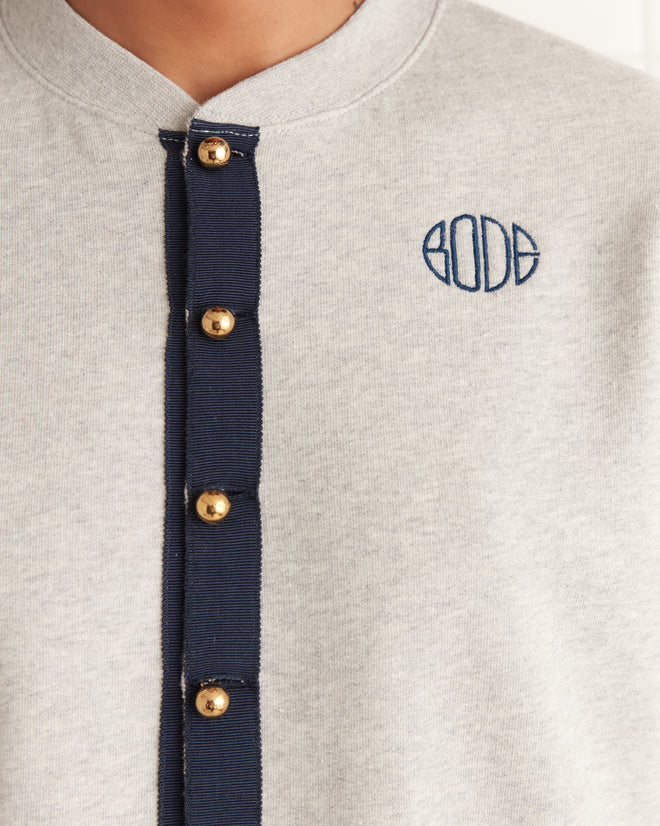 Close-up of a Heathered Cardigan - Grey with gold buttons and a BODE logo embroidered in blue on heathered grey jersey fabric.