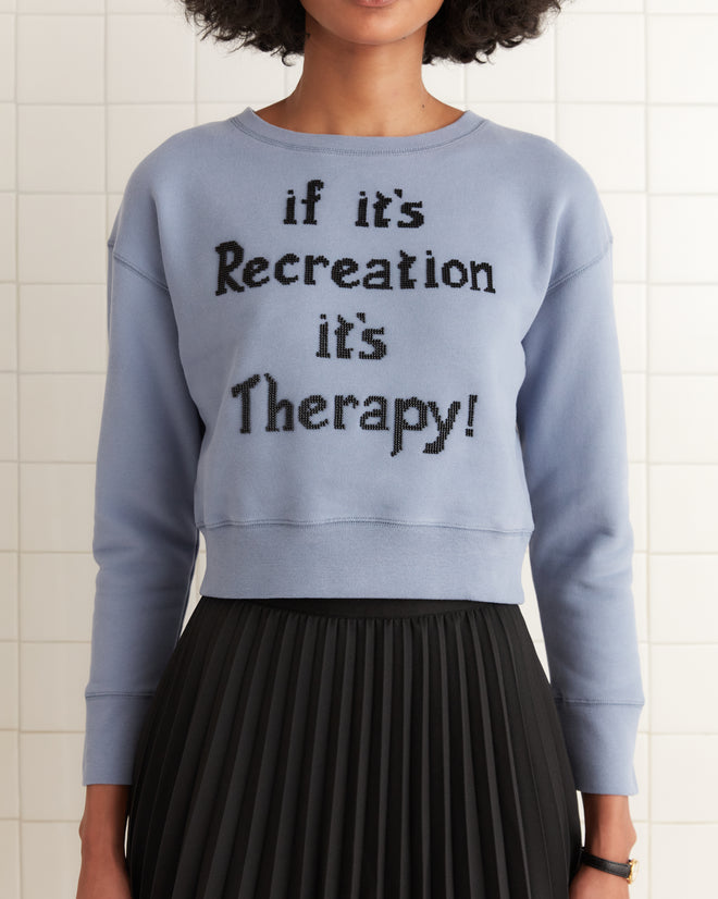 Someone is sporting a cropped lavender Recreation Crewneck, made from 100% cotton and featuring the phrase "if it's Recreation it's Therapy!" combined with a black pleated skirt.