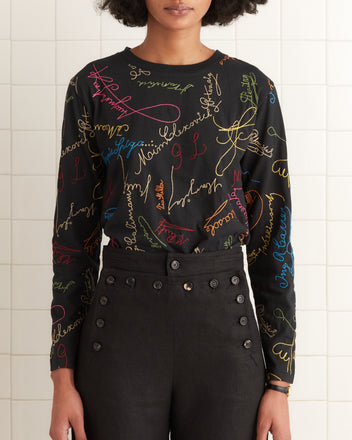 Wearing the Signature Scrawl Long Sleeve Tee with vibrant cursive and high-waisted black pants, a person stands against a tiled wall, exuding cool style. This comfy ensemble, machine washable in cold water, is perfect for those who value both fashion and ease.