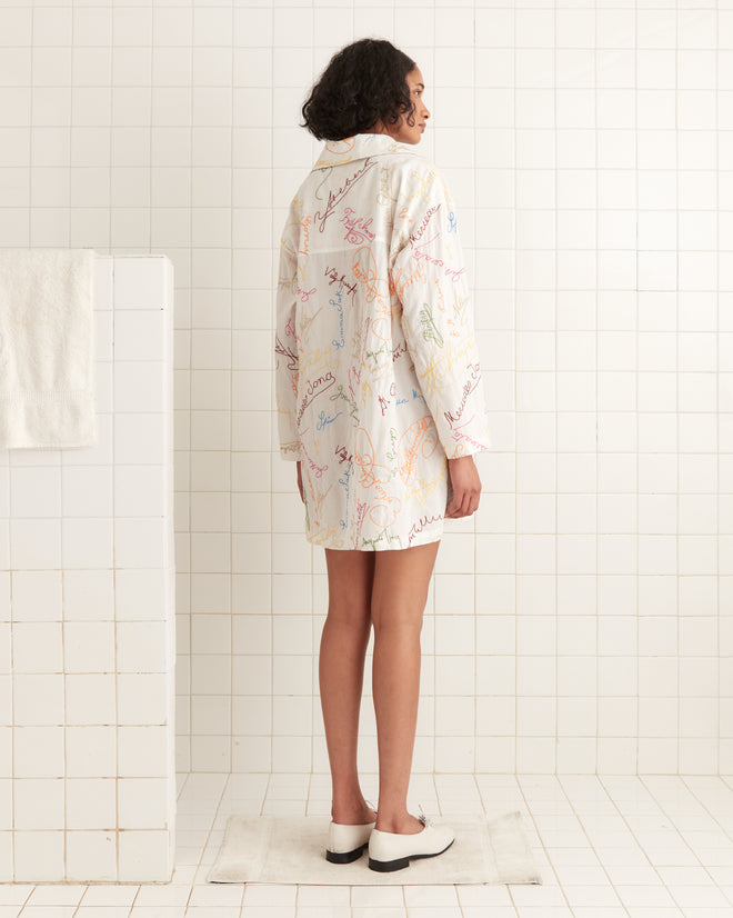 A person with curly hair in a Signature Scrawl Dress stands in a white tiled room, facing away.