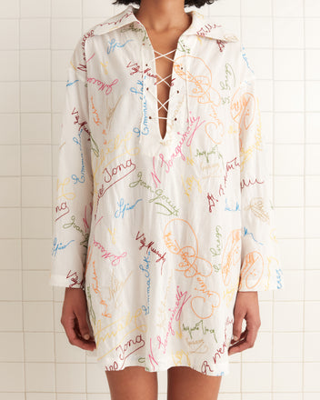 A person is wearing the Signature Scrawl Dress, a white shirt dress made from 100% cotton, featuring colorful embroidered words and a lace-up neckline.