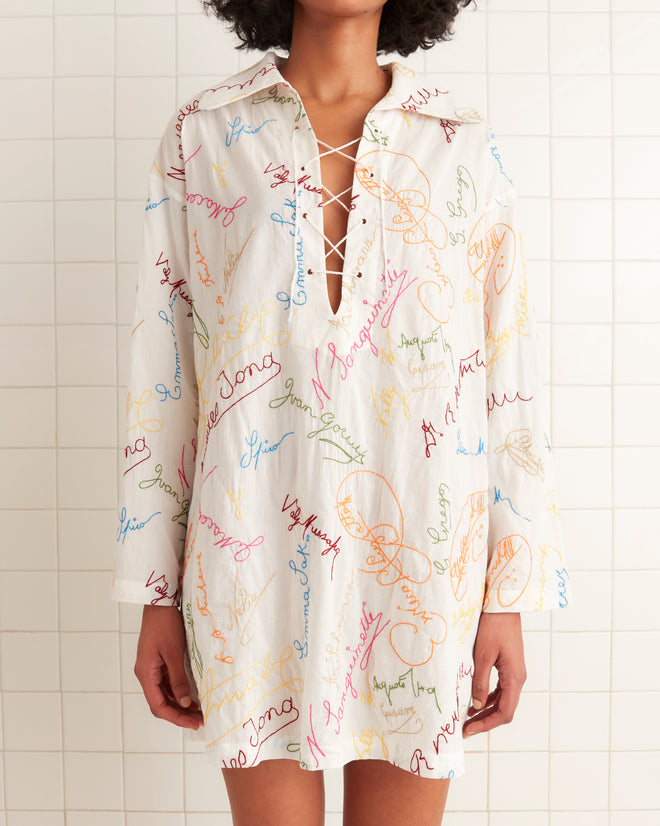 A person wearing the Signature Scrawl Dress, a white dress embellished with colorful, handwritten-style text and featuring a lace-up neckline.