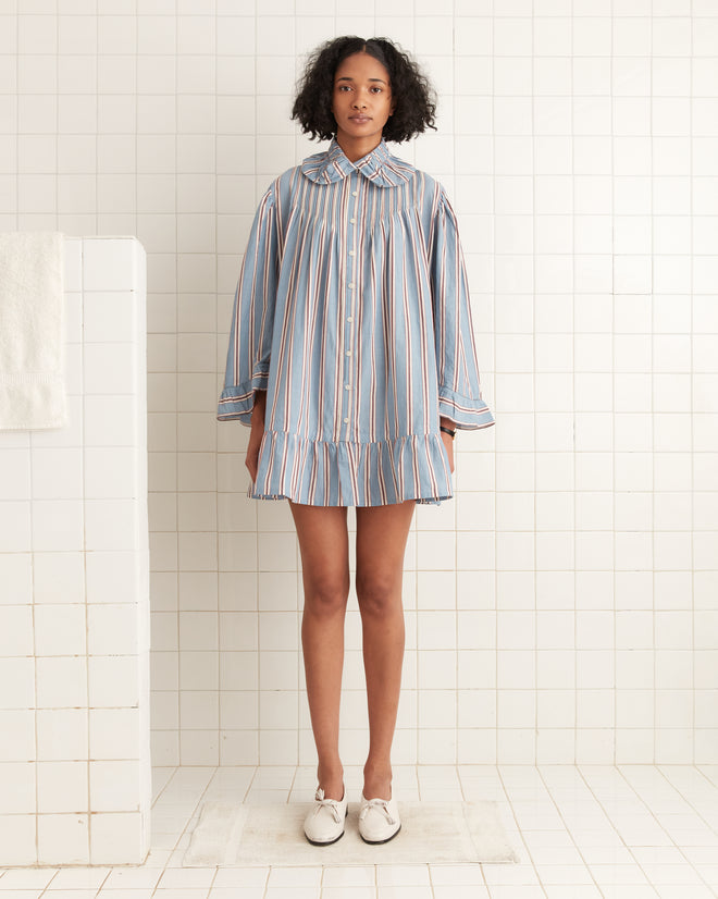 In a tiled room, a person stands in white shoes wearing the Regatta Stripe Dress, an oversized blue-striped shirt made from 100% cotton.