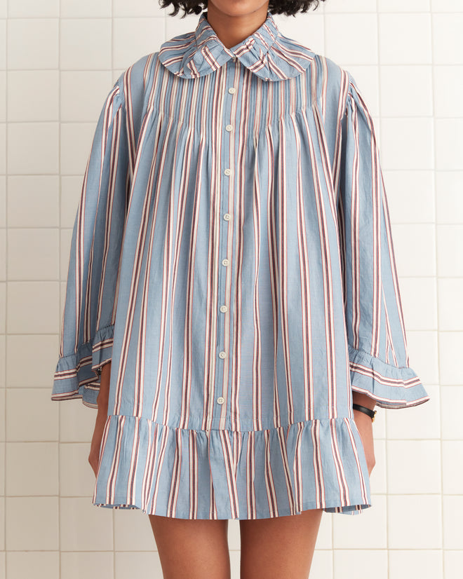 A person wearing the Regatta Stripe Dress, a blue vintage-style piece with a pinstripe pattern, ruffled hem, and collar, stands against a tiled wall, showcasing its timeless elegance and shirting fabric.