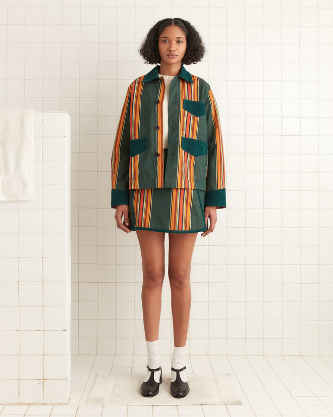 Dressed in a Parakeet Stripe Craft Jacket and skirt set, a person stands in a tiled room, exuding classic workwear elegance with a towel casually draped nearby.