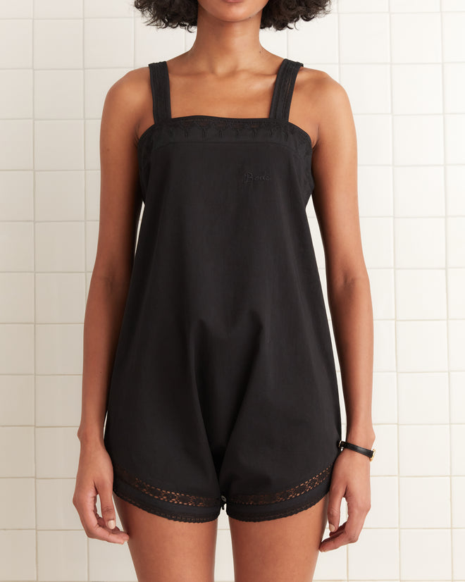Woman wearing the Convertible Teddy - Black, a sleeveless lace romper with delicate trim, standing against a tiled wall.