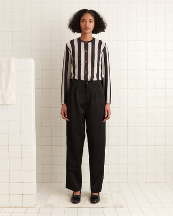 A person in a striped shirt and black pants stands in a white-tiled room, with their outfit subtly enhanced by the gleam of lurex yarn from the Grandstand Cardigan.