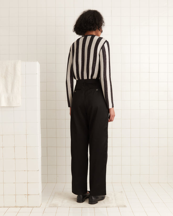 A person with short hair wearing a Grandstand Cardigan with lurex yarn and black pants stands in a tiled room, facing away.
