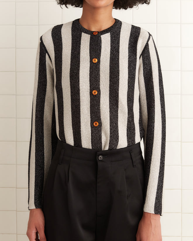 A person stands confidently against a tiled wall, wearing the Grandstand Cardigan featuring black and white stripes with subtle glitter, along with black pants.