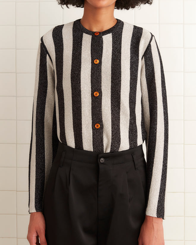 A person wearing the Grandstand Cardigan, featuring black and white knit with glitter lurex yarn stripes and wooden buttons, paired with black pants, stands against a tiled wall.