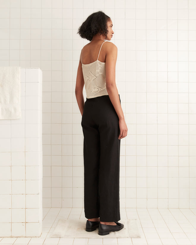 A woman wears the Angel Fish Tank - Cream, a sleeveless knit lace top, paired with black pants as she stands in a white tiled bathroom facing away.