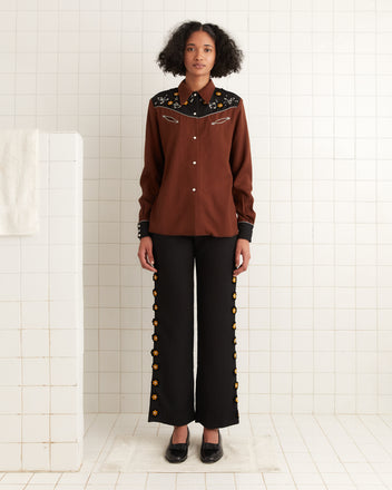 Dressed in the Abilene Western Shirt, embroidered with pearl snap buttons, and black pants with floral accents, a person stands in a tiled room, capturing the essence of a 1960s Western TV show.