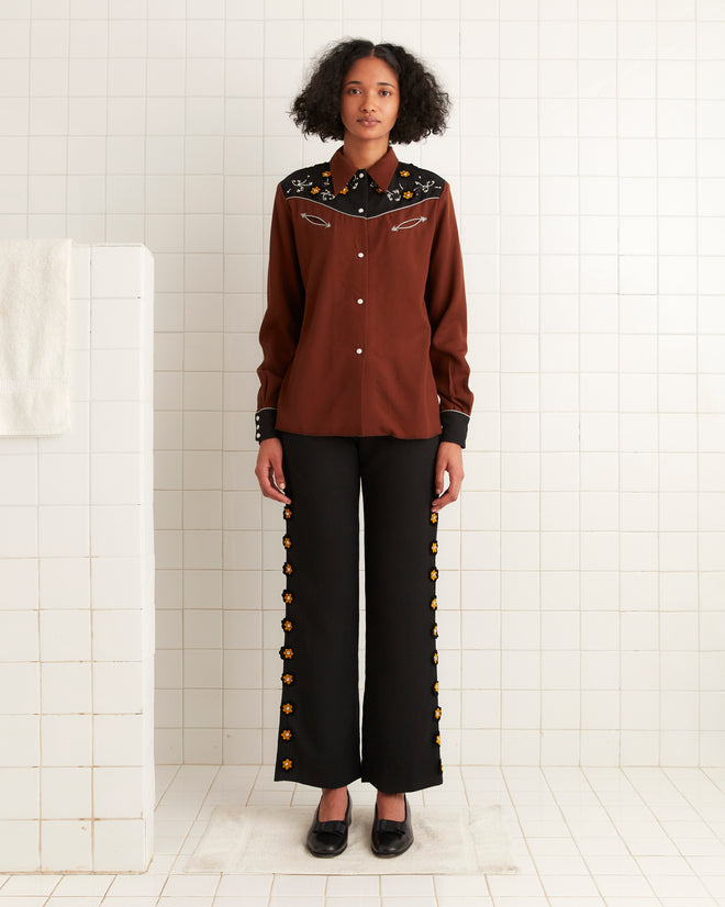 A person wearing the Abilene Western Shirt, featuring floral embroidery and pearl snap buttons, is paired with black pants in a tiled room.