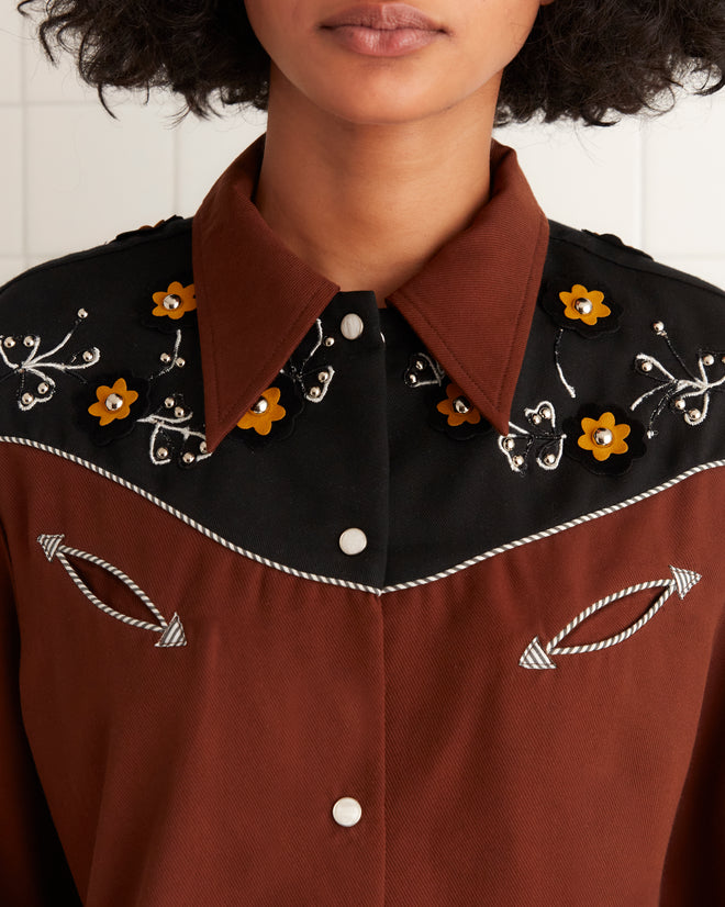 A woman wears the Abilene Western Shirt, featuring brown fabric with floral embroidery and classic pearl snap buttons.