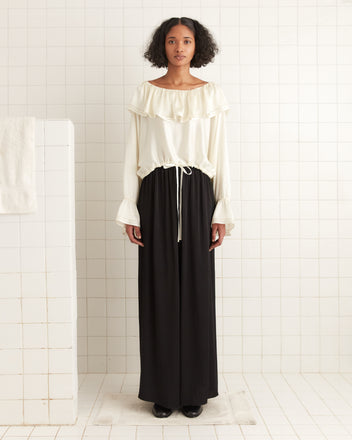 A woman elegantly stands on a white rug in a tiled room, wearing a Satin Claret Blouse in white with a ruffled collar and a black skirt.