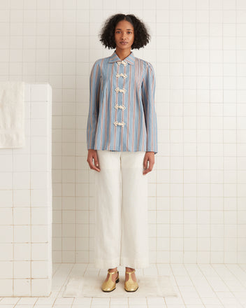 In a tiled room, someone displays the Regatta Stripe Jonas Shirt with pintuck details, paired with white pants and gold shoes, crafting an elegant look.