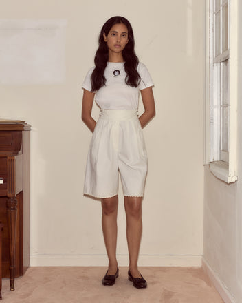 A woman stands indoors by a window on a pale carpet, facing the camera, wearing white Closerie Shorts with a scalloped hem and a matching t-shirt.