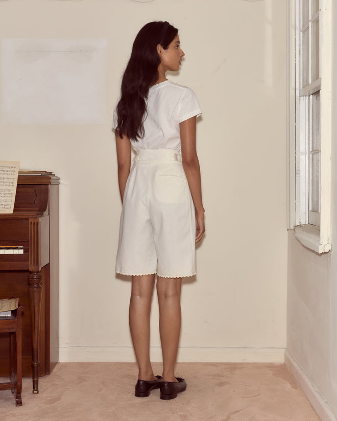 A woman stands facing away in a sunlit room near a piano and sheet music, wearing the Closerie Shorts—an ivory embroidered piece paired with a white outfit.