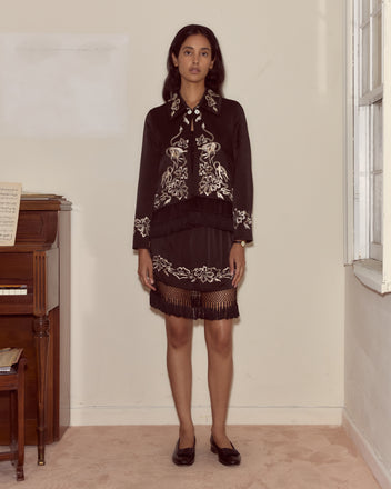 Woman in a black Phantasm Fringe Skirt stands indoors by a piano and window, looking at the camera.