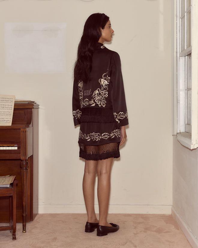 A woman in a black Phantasm Fringe Skirt stands by a piano, facing away near a window in a softly lit room.